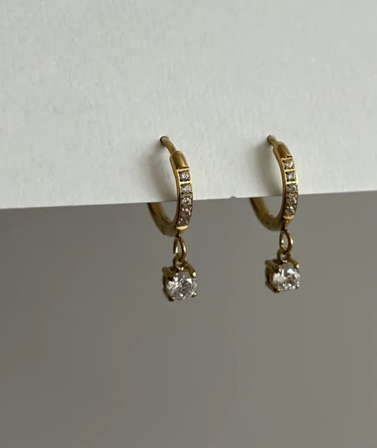 A pair of gold-plated huggie hoop earrings with dangling crystal charms, shown on a smartphone shopping screen with the text 'AMOUR CRYSTAL EARRINGS-18K GOLD PLATED' and a price of Rs. 749.